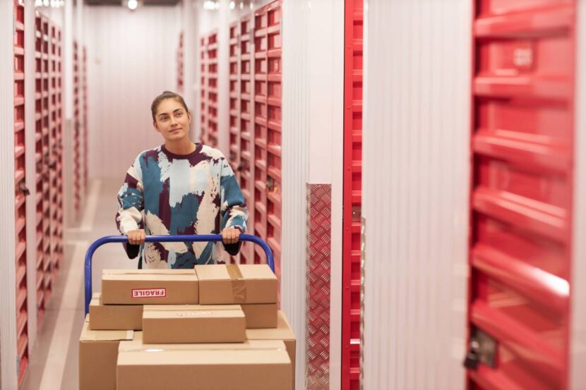 What is Self-Storage? How Self-Storage Can Ease Your Life Smiling woman pushing big cart with cardboard boxes along corridor of self-storage